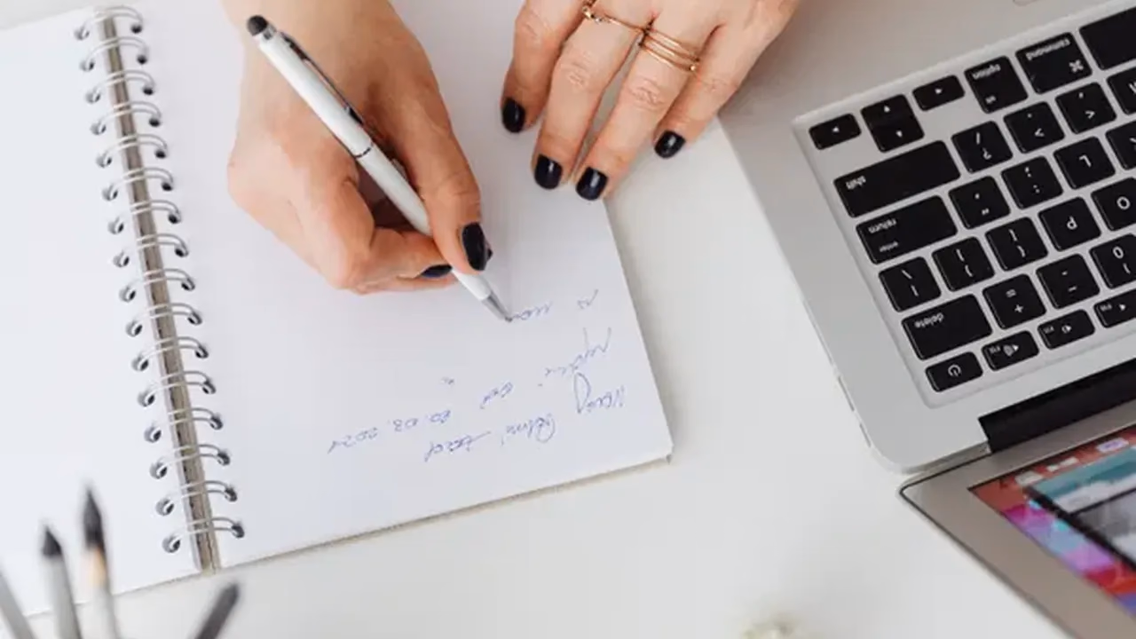 Hands writing post notes in a spiral notebook beside a laptop